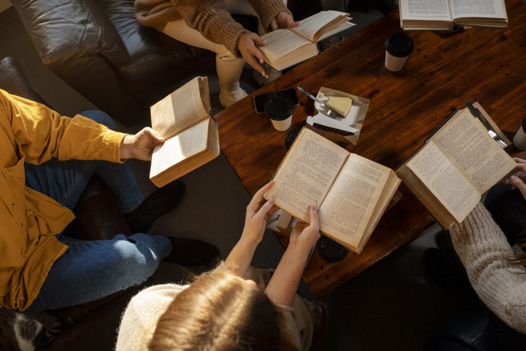 Foto tirada do ângulo de cima, focando nos livros que as 5 pessoas ao redor da mesa do clube de leitura estão lendo