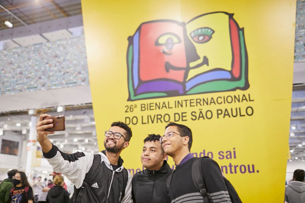 Três homens tirando uma selfie em frente ao banner da Bienal do Livro.